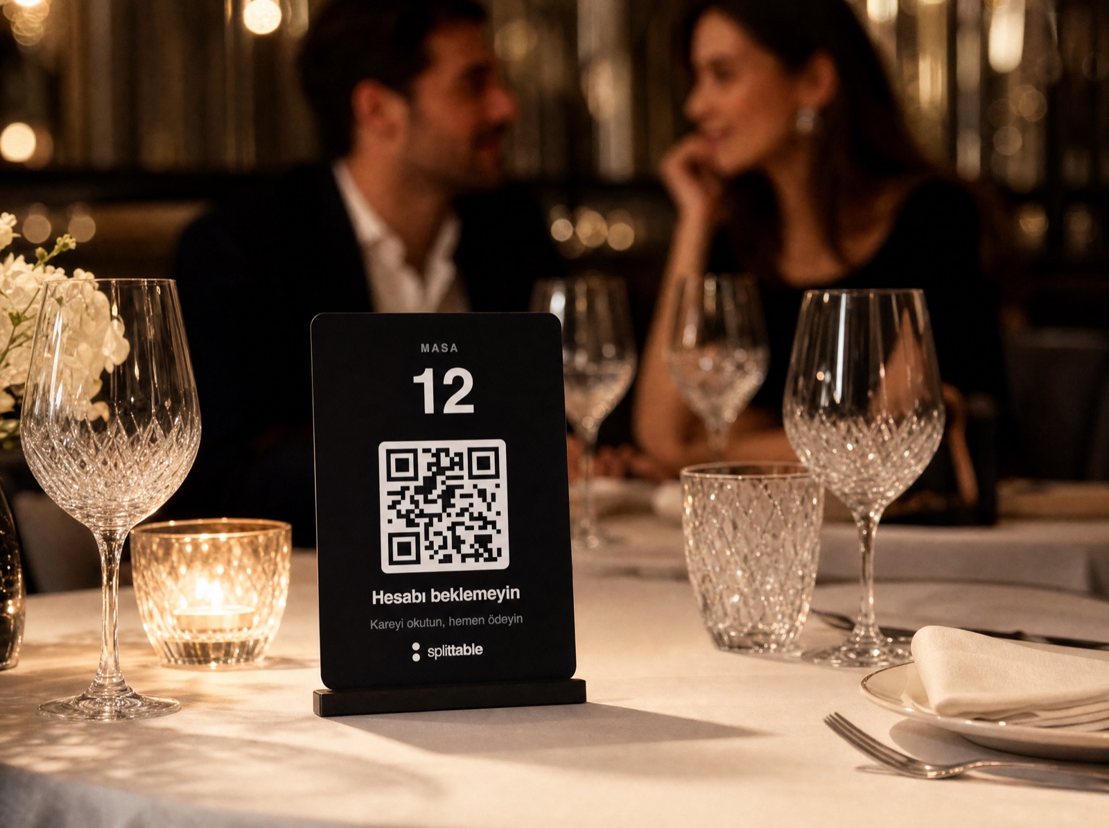 Beyaz örtülü fine dining masasında duran siyah akrilik splittable standı; masa numarası ve karekod bir arada, arkada bir çift oturuyor.